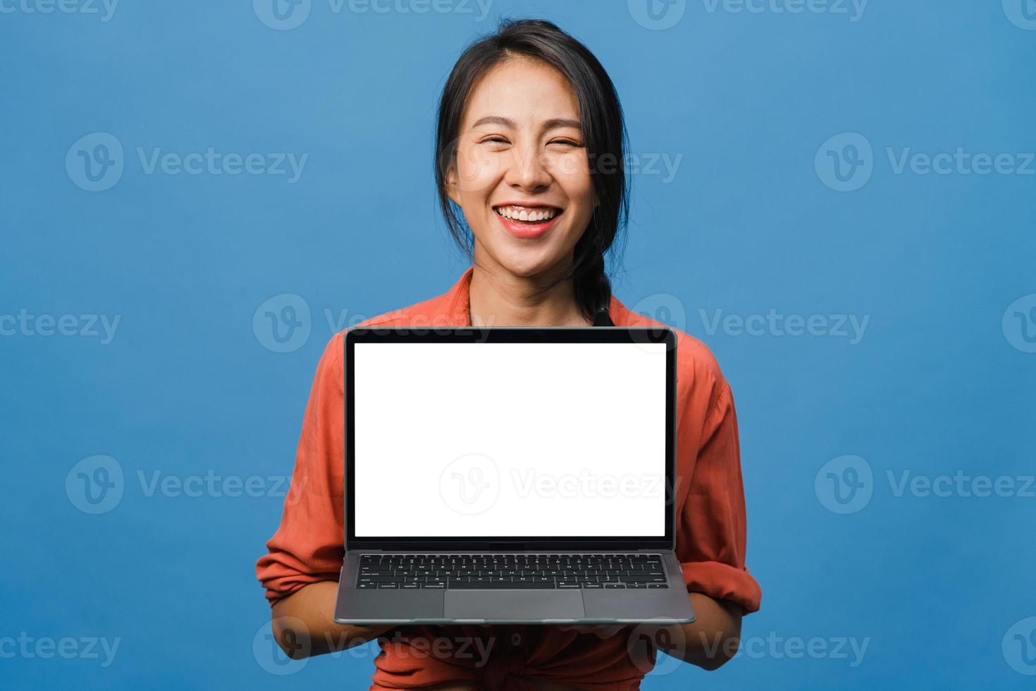 Joven asiática muestra una pantalla de computadora portátil vacía con expresión positiva, sonríe ampliamente, vestida con ropa casual sintiendo felicidad aislada sobre fondo azul. computadora con pantalla blanca en mano femenina. foto