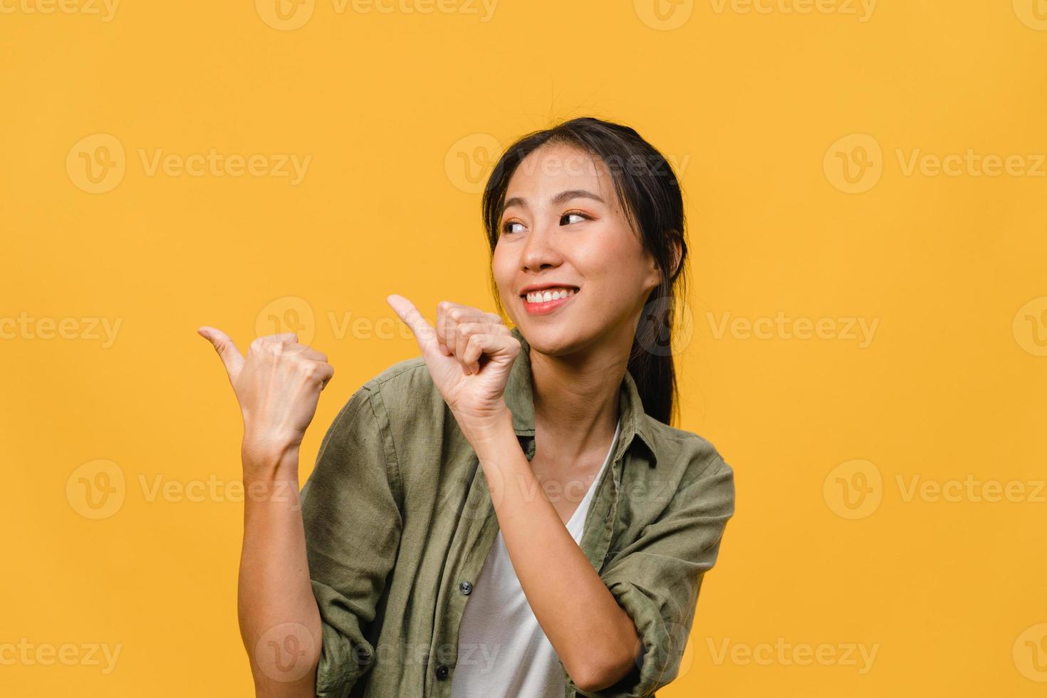 Portrait of young Asian lady smiling with cheerful expression, shows something amazing at blank space in casual clothing and standing isolated over yellow background. Facial expression concept. photo
