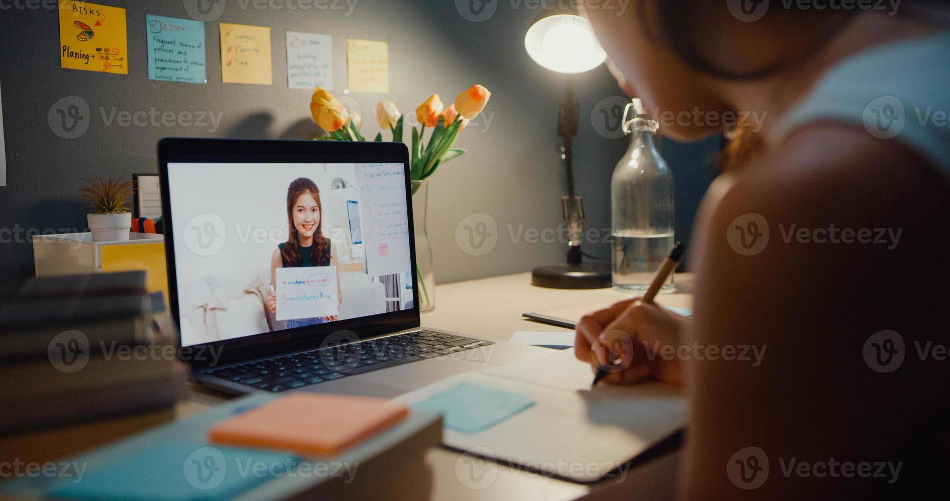 Young Asia teen girl student distance learning lesson with online teacher and study on laptop screen in living room at home night. Remotely, Social distancing, quarantine for corona virus prevention. photo