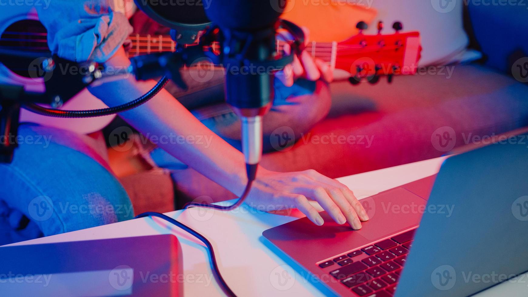 blogger de niña asiática feliz toca la guitarra y usa el micrófono, canta una canción, graba música, mezclador de sonido en la computadora portátil en la moderna sala de estar, estudio en casa por la noche. creador de contenido musical, tutorial, concepto de transmisión. foto