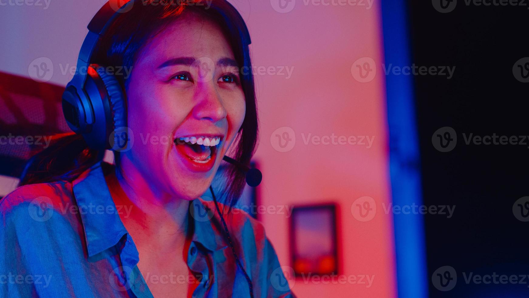 Close-up face happy asia professional girl gamer wear headphone competition play video game neon light computer in living room at home. Esport streaming game online, Home quarantine activity concept. photo