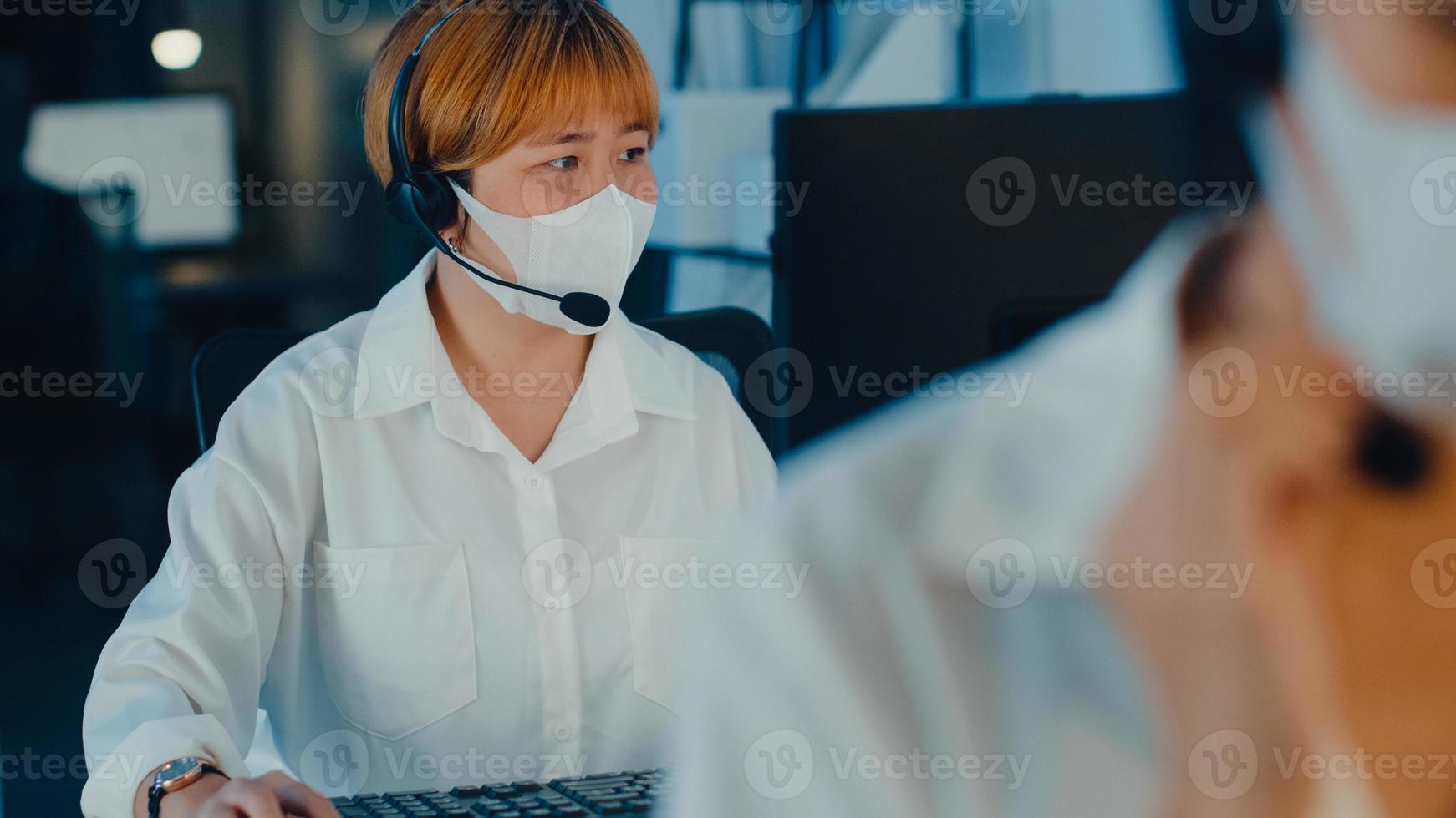 Millennial Asia young call center team or customer support service executive wearing face mask prevent covid-19 using computer and microphone headset working technical support in late night office. photo