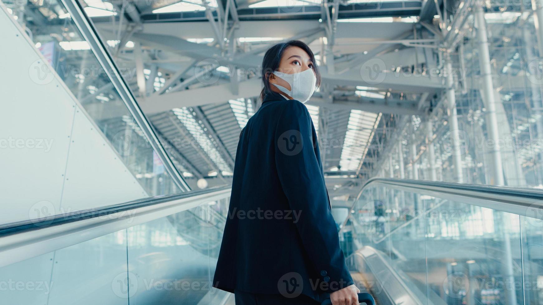 Asian business girl wear face mask drag luggage stand on escalator look around walk to terminal at international airport. Business commuter covid pandemic, Business travel social distancing concept. photo