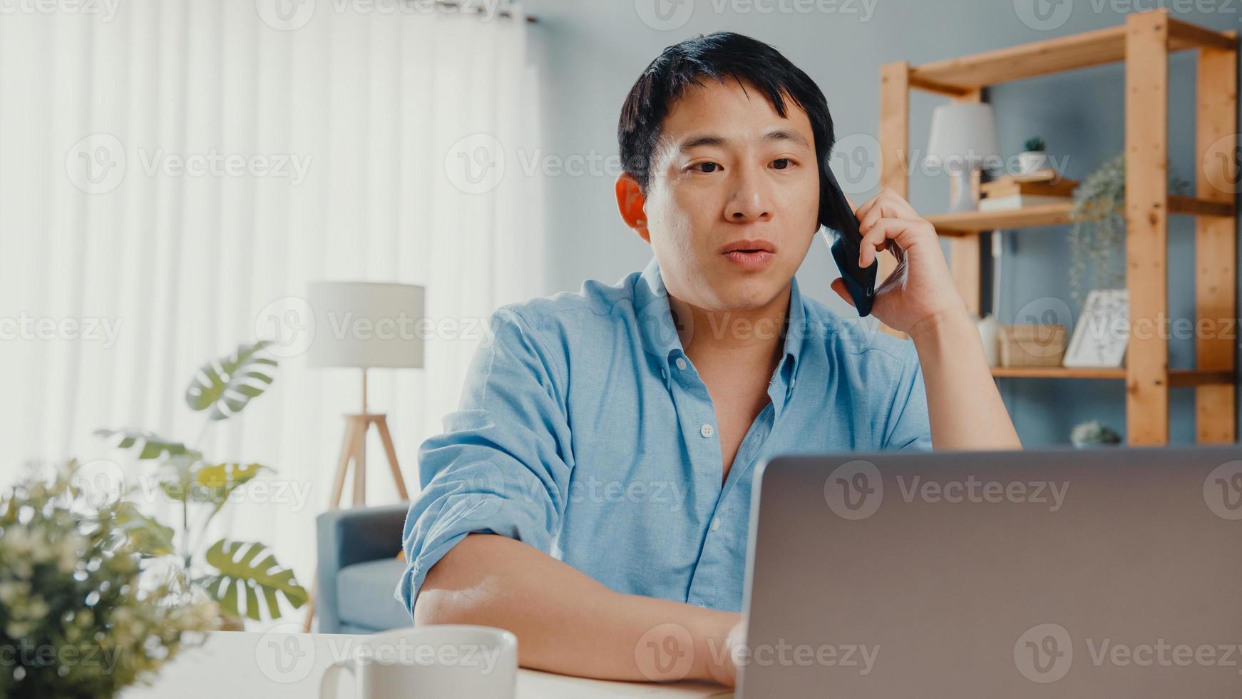 freelance chico asiático ropa casual usando laptop hablar por teléfono celular en la sala de estar en casa. trabajo desde casa, trabajo a distancia, educación a distancia, distanciamiento social, cuarentena para la prevención del coronavirus. foto