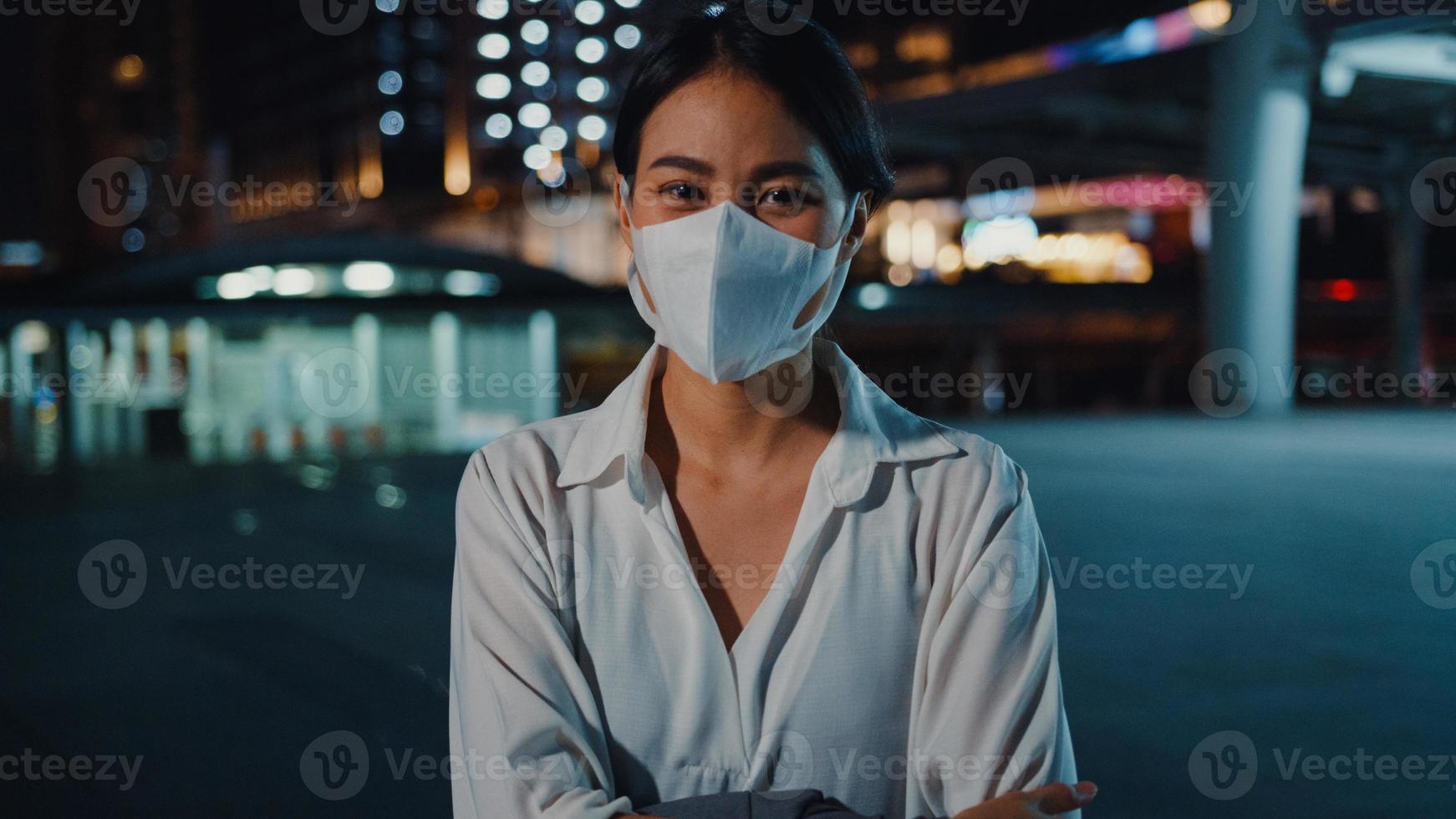 Successful young Asia businesswoman in fashion office clothes wear medical face mask smiling and looking at camera while happy stand alone outdoors in urban modern city night. Business on go concept. photo