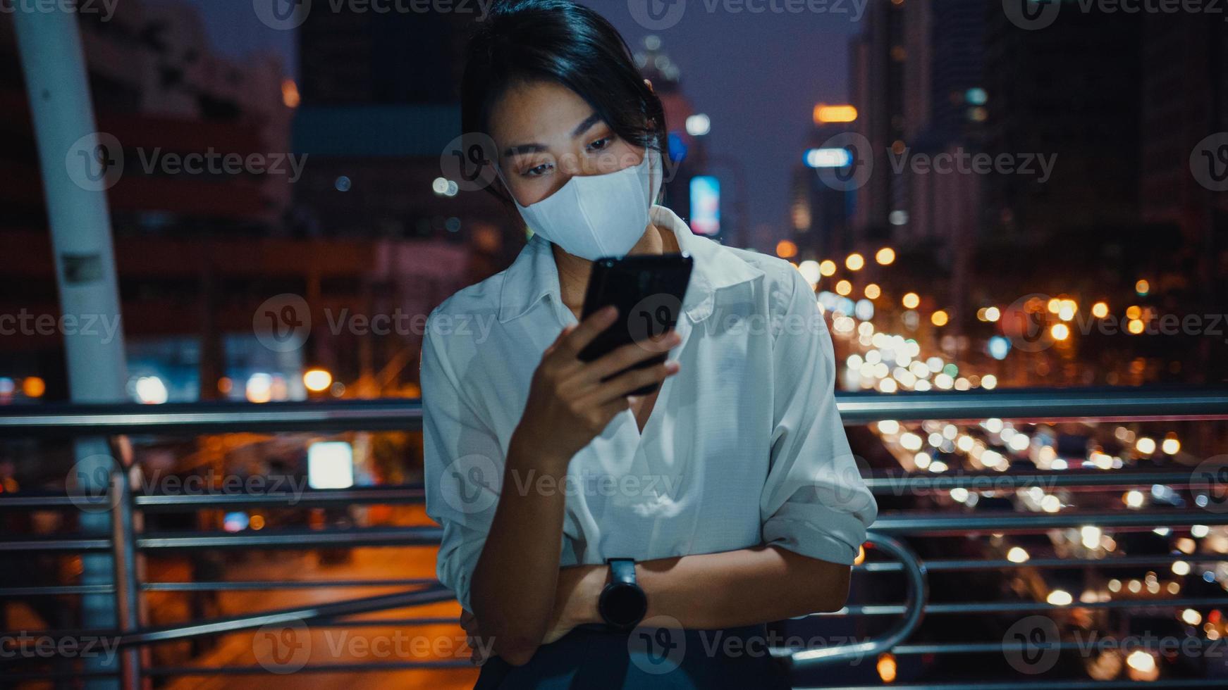 Joven empresaria asiática en ropa de moda con mascarilla usando un teléfono inteligente escribiendo un mensaje de texto mientras está parado al aire libre en la ciudad urbana por la noche. distanciamiento social para evitar la propagación del concepto covid-19. foto