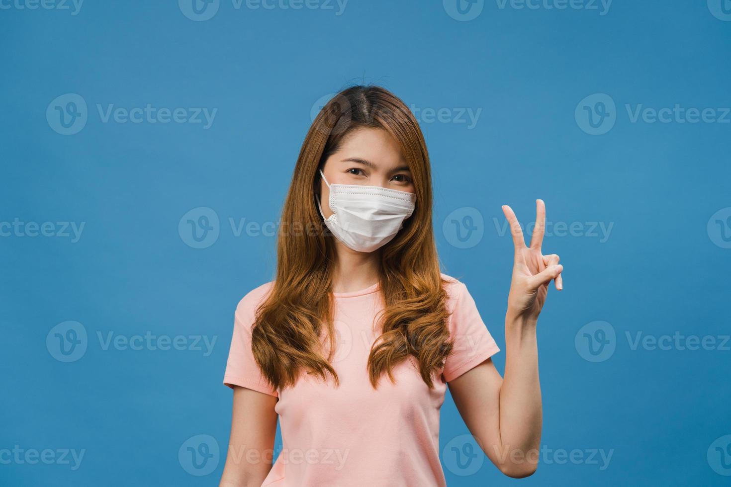 Young Asia girl wearing medical face mask showing peace sign, encourage with dressed in casual cloth and looking at camera isolated on blue background. Social distancing, quarantine for corona virus. photo