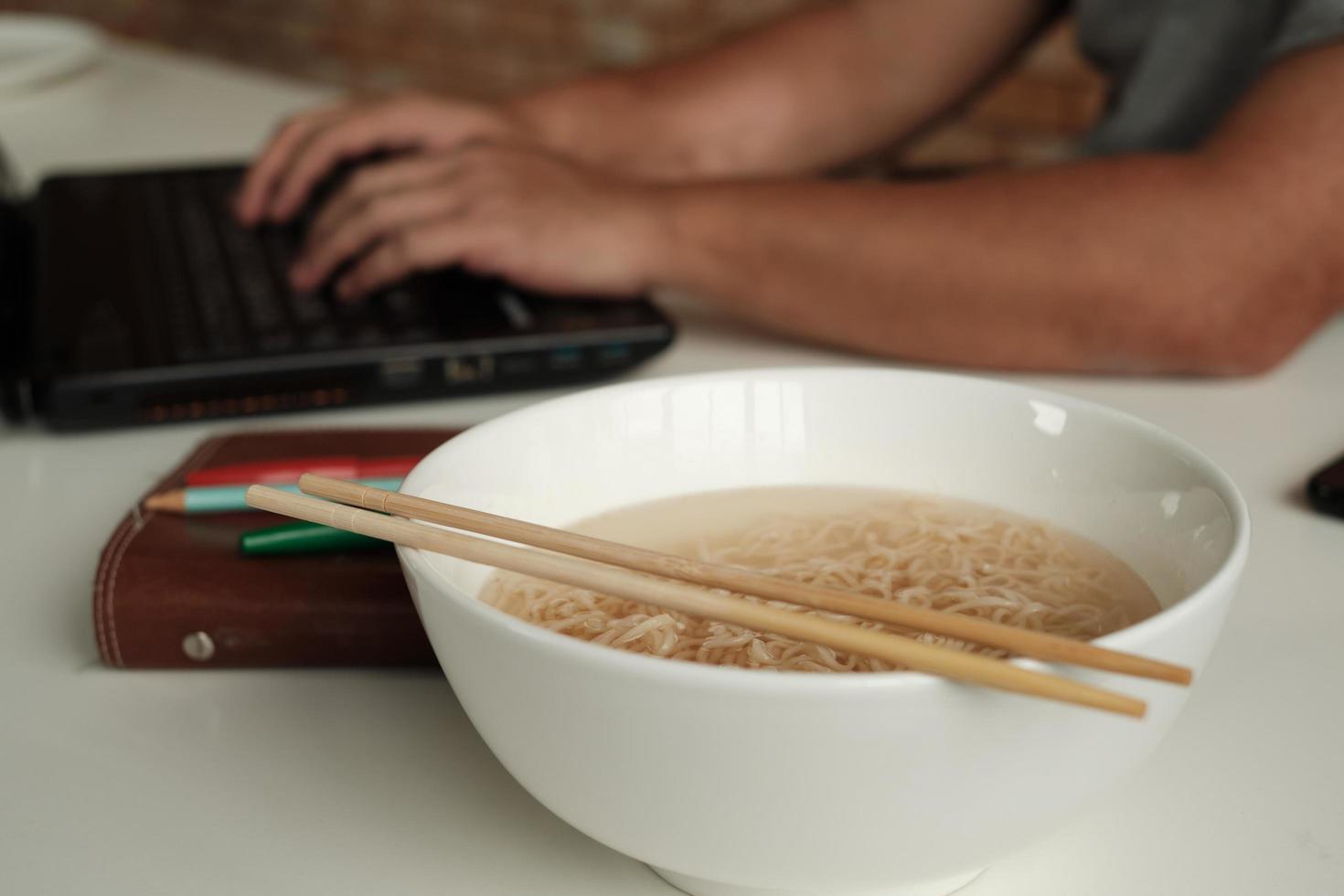 Thai male worker busy working with laptop, use chopsticks to hastily eat instant noodles during office lunch's break, because quick, tasty, and cheap. Over time Asian fast food, unhealthy lifestyle. photo