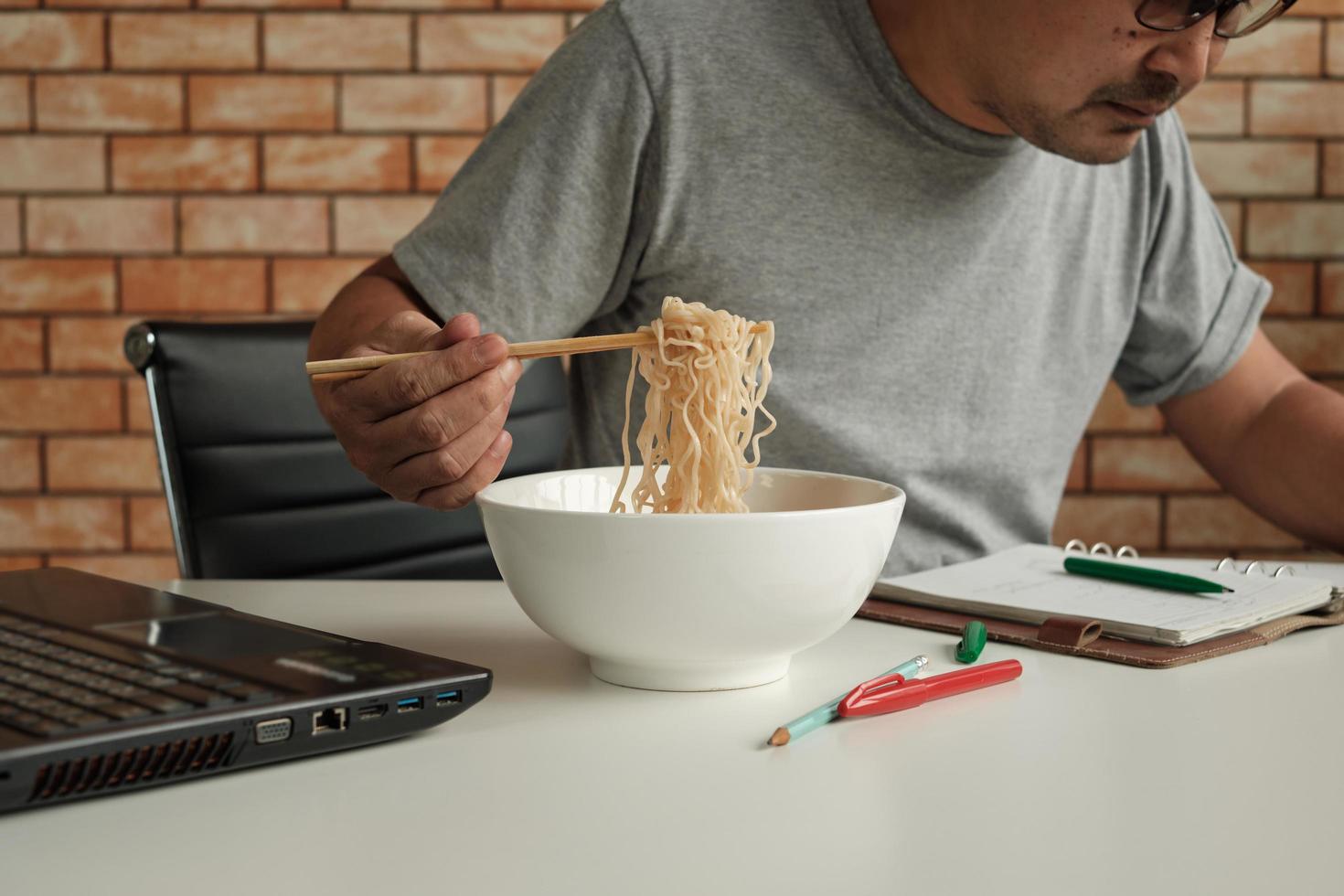 Trabajador tailandés ocupado trabajando con una computadora portátil, use palillos para comer apresuradamente fideos instantáneos durante el almuerzo de la oficina, porque es rápido, sabroso y barato. con el tiempo comida rápida asiática, estilo de vida poco saludable. foto