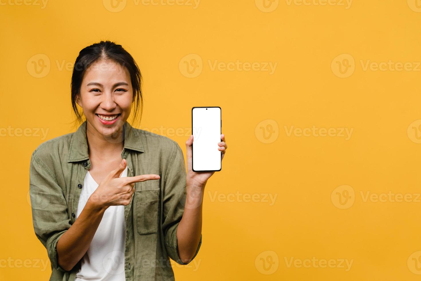 Joven asiática muestra la pantalla vacía del teléfono inteligente con expresión positiva, sonríe ampliamente, vestida con ropa casual sintiendo felicidad sobre fondo amarillo. teléfono móvil con pantalla en blanco en mano femenina. foto