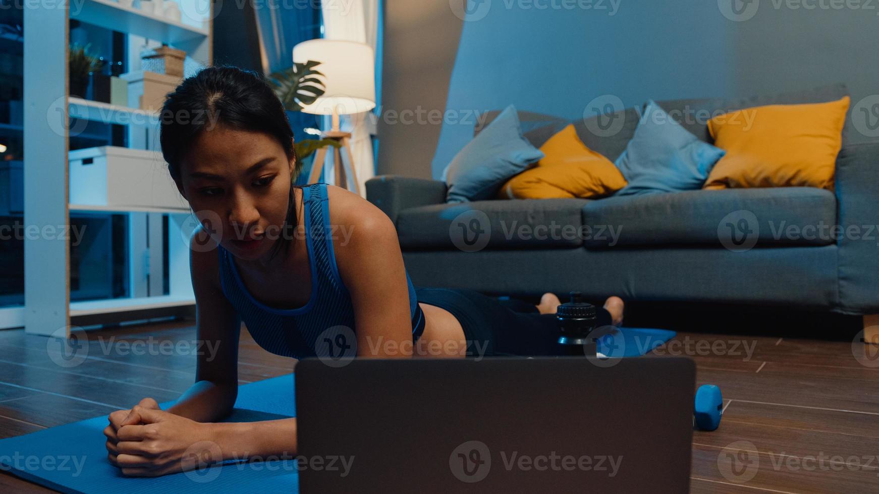 Jovencita asiática en ejercicios de ropa deportiva haciendo ejercicio y usando la computadora portátil para ver un video tutorial de yoga en la noche en casa. entrenamiento a distancia con entrenador personal, distancia social, concepto de educación en línea. foto