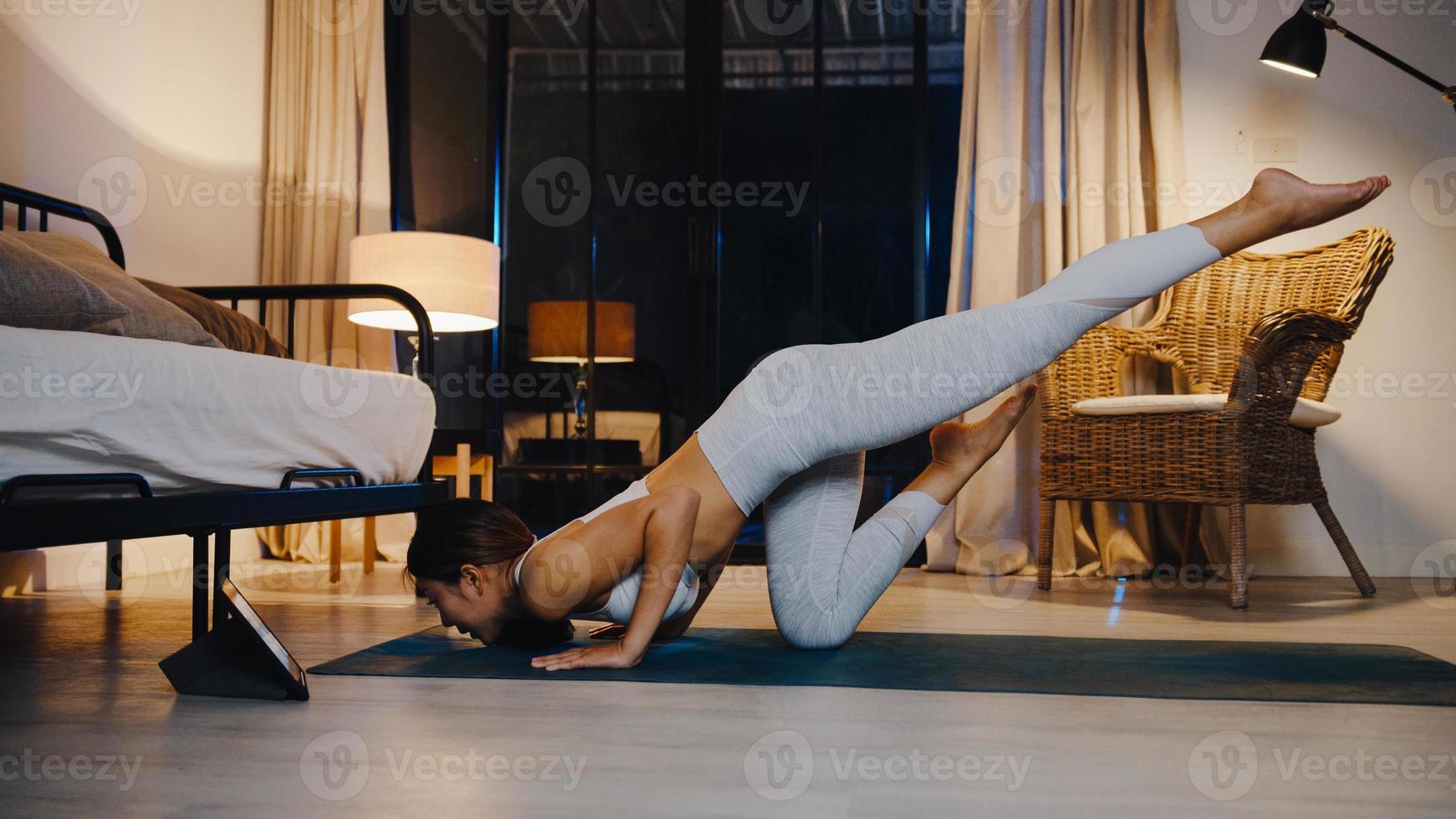 joven asiática en ejercicios de ropa deportiva haciendo ejercicio y usando la tableta para ver el video tutorial de yoga en la noche en casa. entrenamiento a distancia con entrenador personal, distancia social, concepto de educación en línea. foto