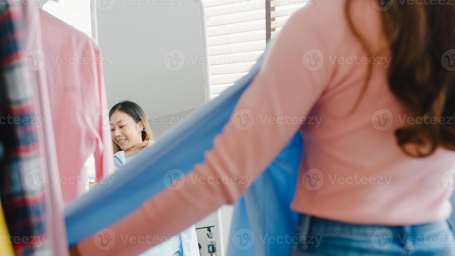 Bella dama atractiva de Asia eligiendo ropa en perchero vestirse mirando a sí misma en el espejo en el dormitorio en casa. chica piensa qué ponerse camisa casual. las mujeres de estilo de vida se relajan en el concepto de hogar. foto