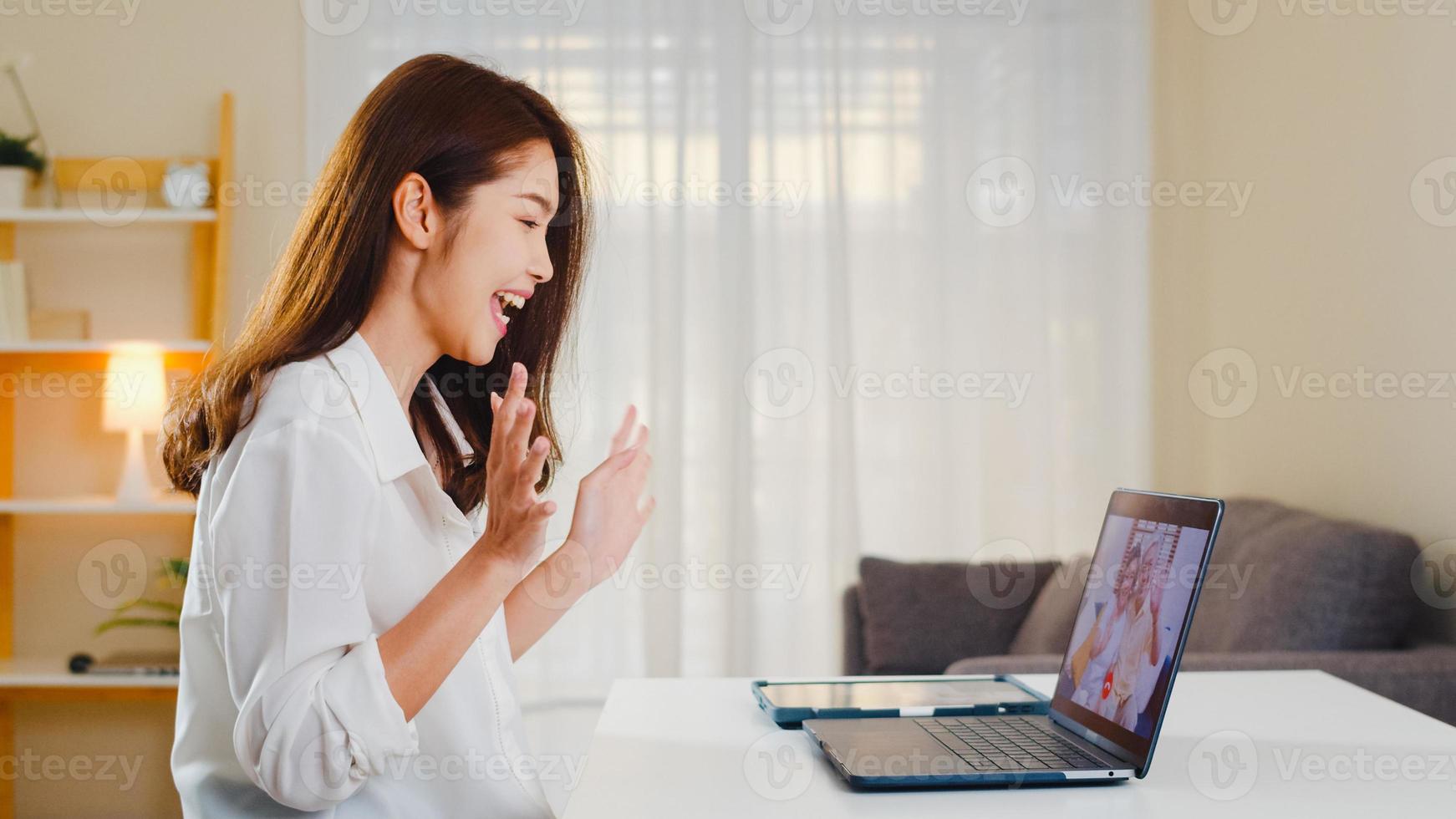 Mujer de negocios asiática joven que usa videollamada portátil hablando con papá y mamá de la familia mientras trabaja desde casa en la sala de estar. autoaislamiento, distanciamiento social, cuarentena para la prevención del coronavirus. foto