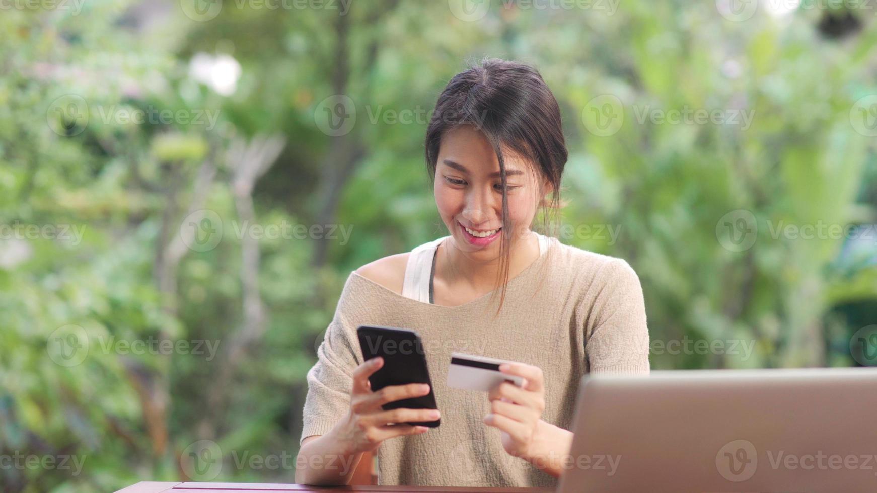 Mujer asiática con teléfono móvil y comercio electrónico de compras con tarjeta de crédito, mujer relajarse sintiéndose feliz de compras en línea sentado en la mesa en el jardín por la mañana. las mujeres de estilo de vida se relajan en el concepto de hogar. foto