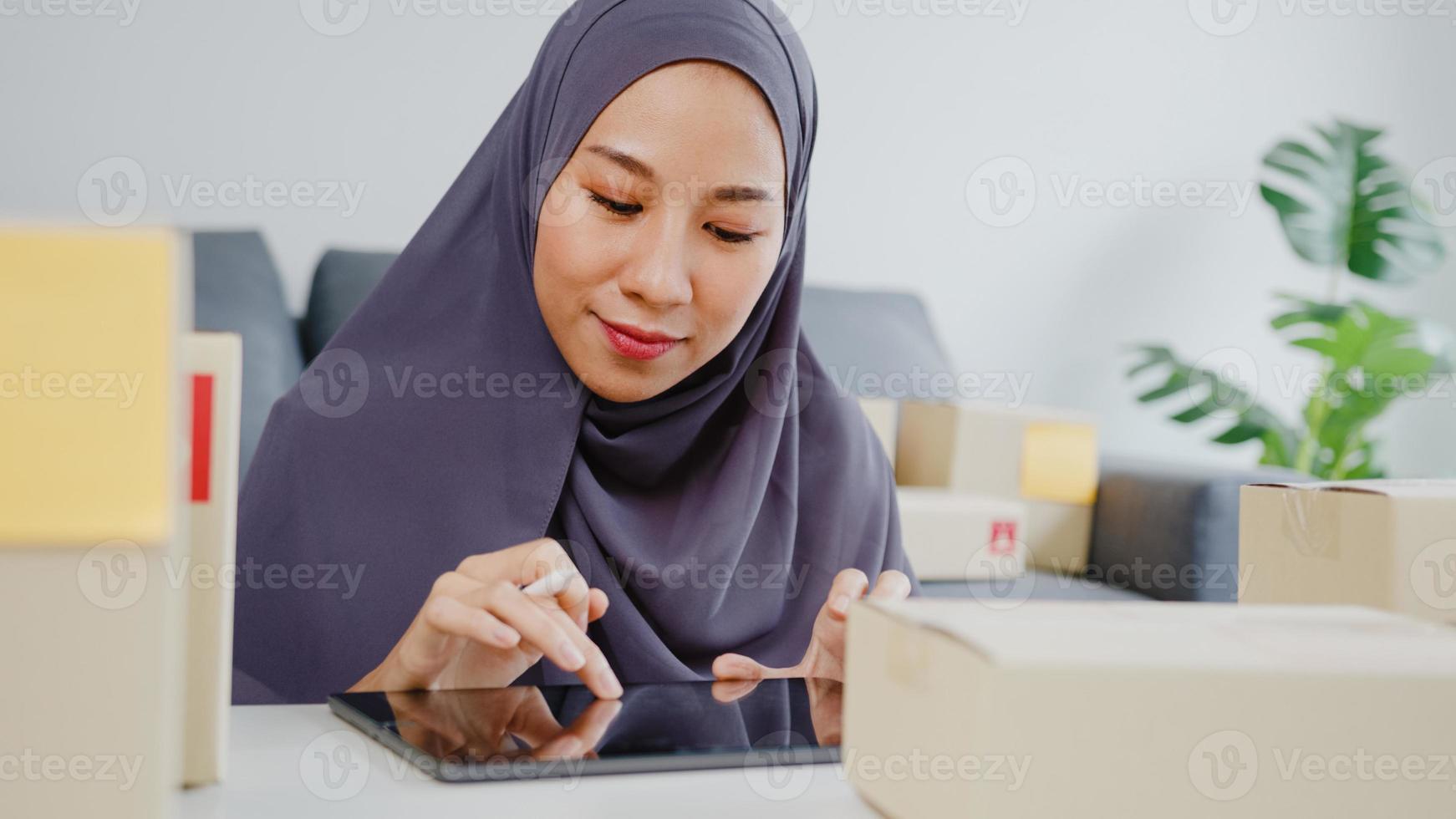 Young Asia muslim businesswoman check product purchase order on stock and save to tablet computer work at home office. Small business owner, online market delivery, lifestyle freelance concept. photo