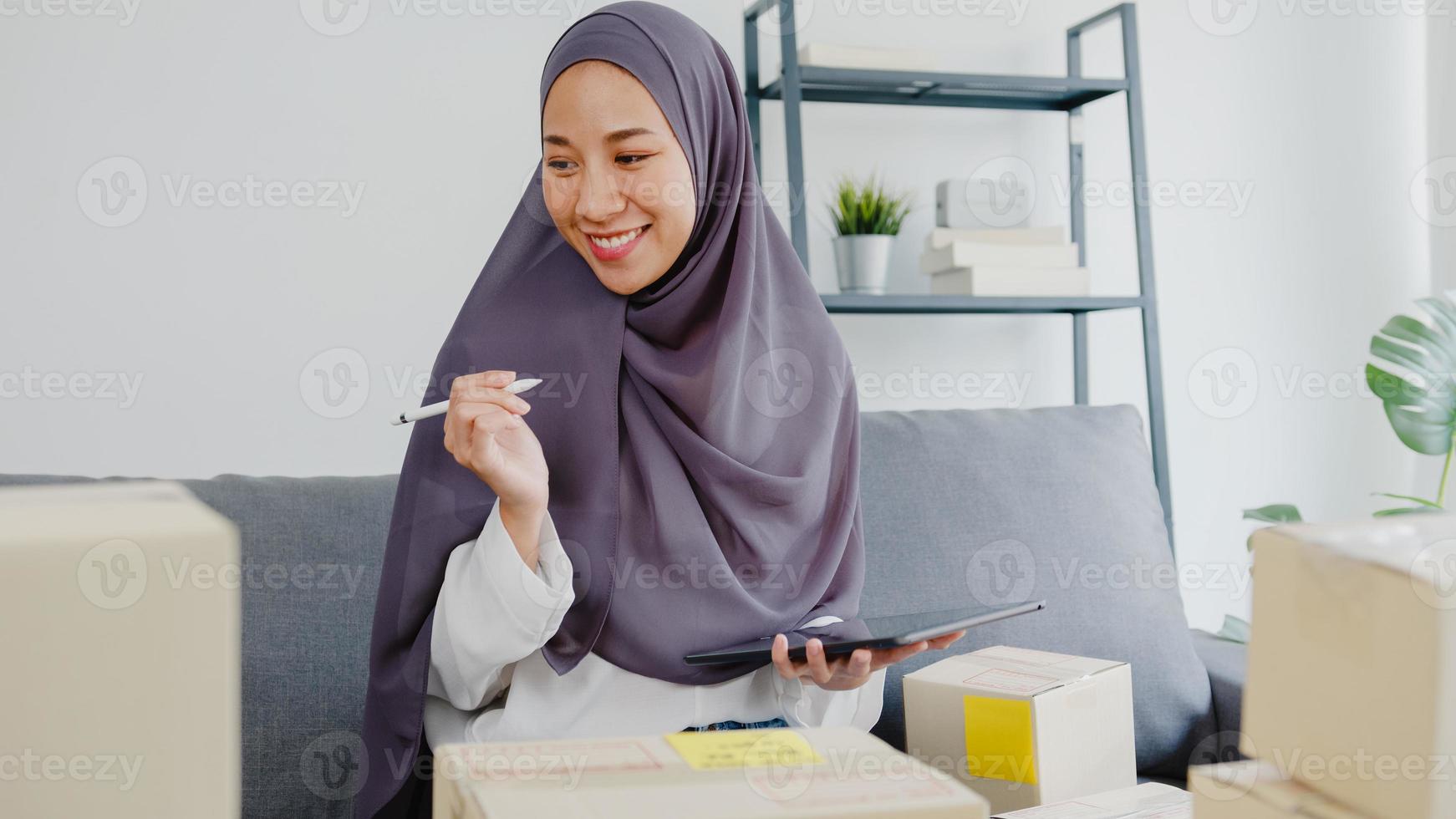 Young Asia muslim businesswoman check product purchase order on stock and save to tablet computer work at home office. Small business owner, online market delivery, lifestyle freelance concept. photo