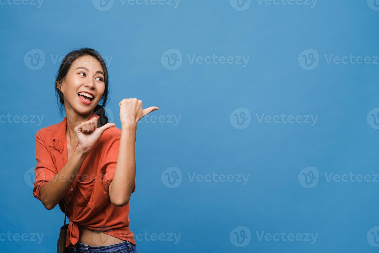 Portrait of young Asian lady smiling with cheerful expression, shows something amazing at blank space in casual clothing and standing isolated over blue background. Facial expression concept. photo