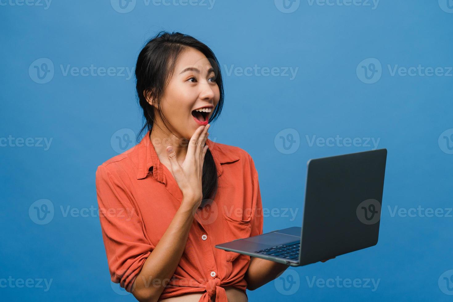 Jovencita asiática usando laptop con expresión positiva, sonríe ampliamente, vestida con ropa casual sintiendo felicidad y parada aislada sobre fondo azul. feliz adorable mujer alegre se regocija con el éxito. foto