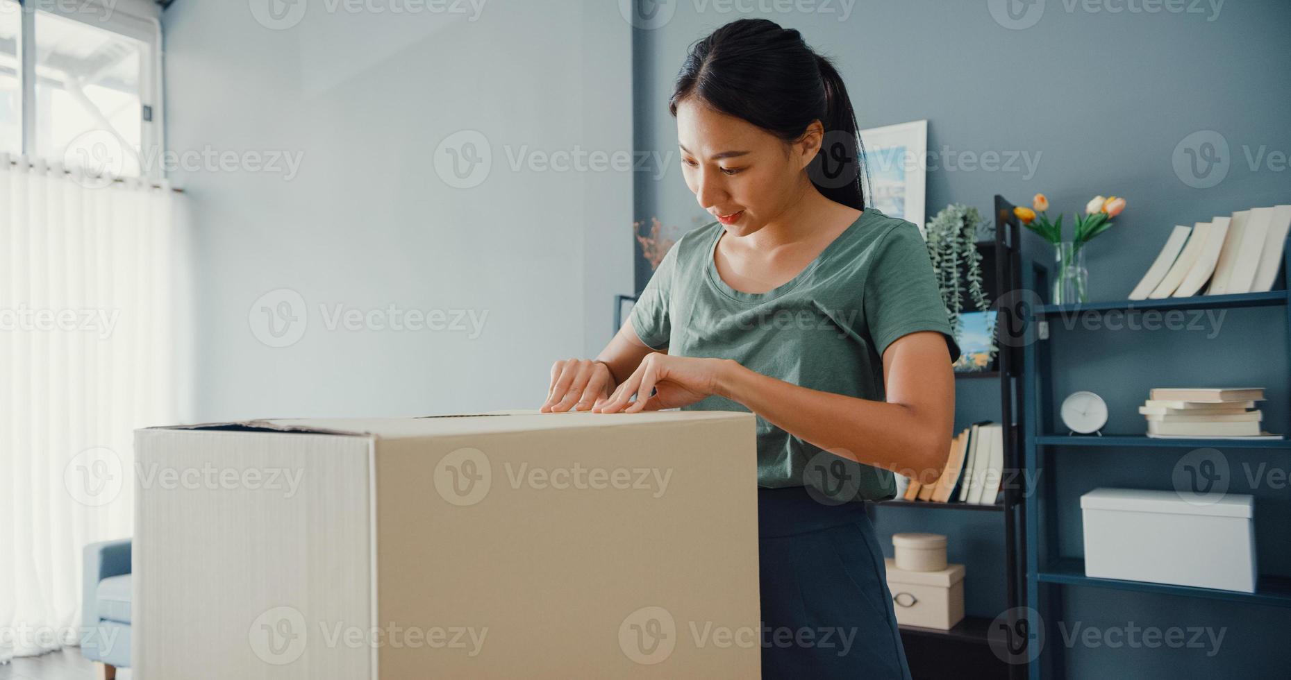 Happy beautiful Asia lady unboxing cardboard delivery package from online marketplace in living room at house. Satisfied shopper in internet unboxing goods, Online shopping and delivery concept. photo