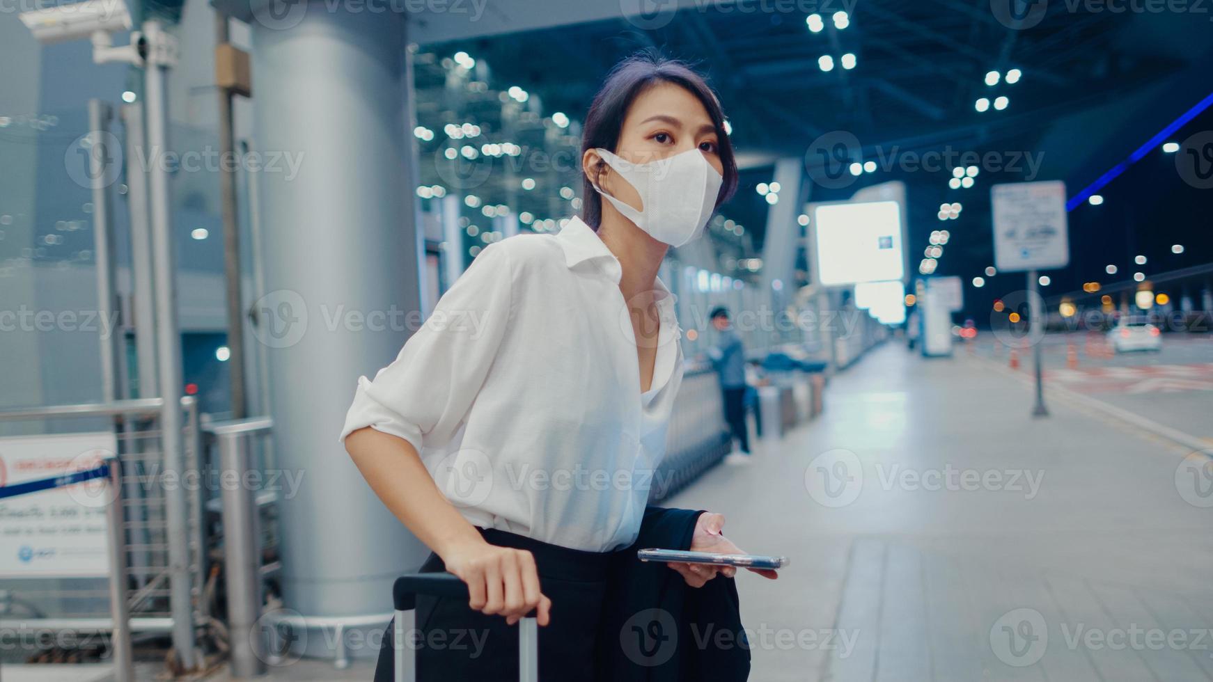 Asian business girl arrive destination wear face mask stand outside look smart phone wait car terminal at domestic airport. Business commuter covid pandemic, Business travel social distancing concept. photo