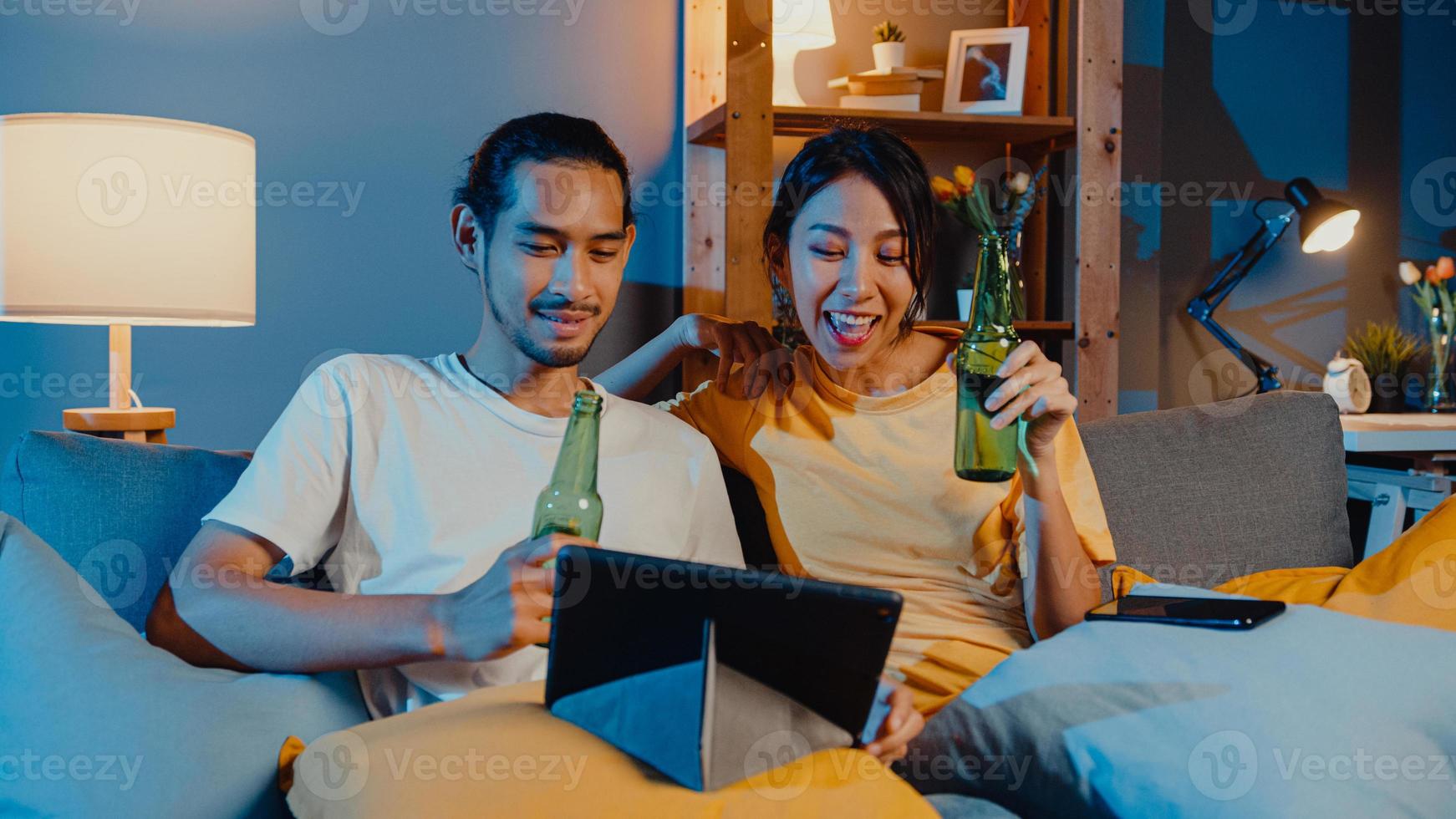 Happy young asian couple enjoy night party event online sit couch use tablet video call with friends toast drink beer via video call online in living room at home, Social distancing concept. photo