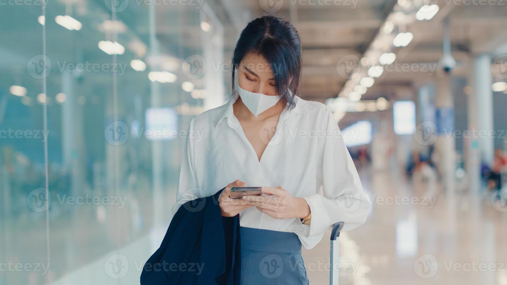 Asian business girl use smart phone for check in boarding pass walk with luggage to terminal at domestic flight at airport. Business commuter covid pandemic, Business travel social distancing concept. photo