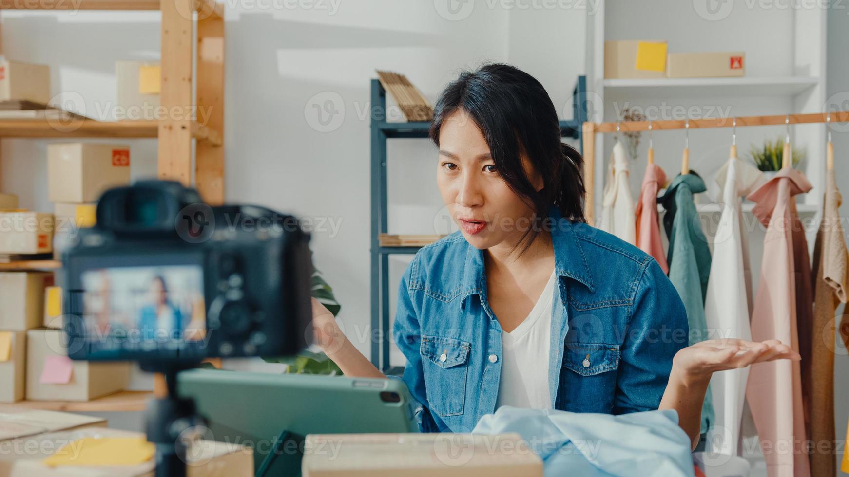 joven diseñadora de moda asiática que usa un teléfono móvil y recibe una orden de compra y muestra la ropa, grabando video en vivo en línea con la cámara. propietario de una pequeña empresa, concepto de entrega de mercado en línea. foto