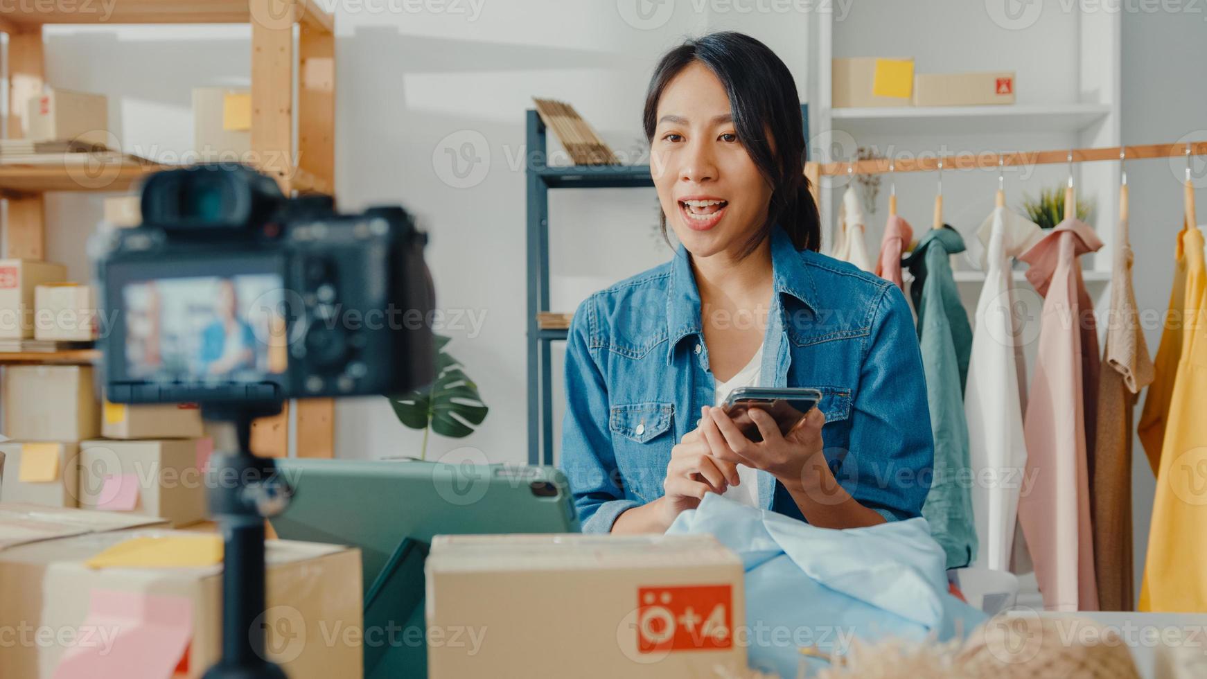 joven diseñadora de moda asiática que usa un teléfono móvil y recibe una orden de compra y muestra la ropa, grabando video en vivo en línea con la cámara. propietario de una pequeña empresa, concepto de entrega de mercado en línea. foto
