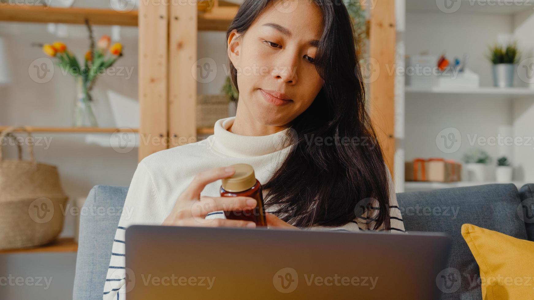 mujer asiática joven enferma mantenga la medicina sentarse en la videollamada del sofá con la computadora portátil consulte con el médico en casa. chica toma medicamentos después de la orden del médico, cuarentena en la casa, concepto de coronavirus de distanciamiento social. foto