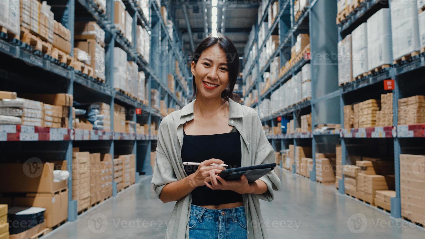 Portrait of attractive young Asia businesswoman manager smiling charmingly looking at camera hold digital tablet stand in retail shopping center. Distribution, Logistics, Packages ready for shipment. photo