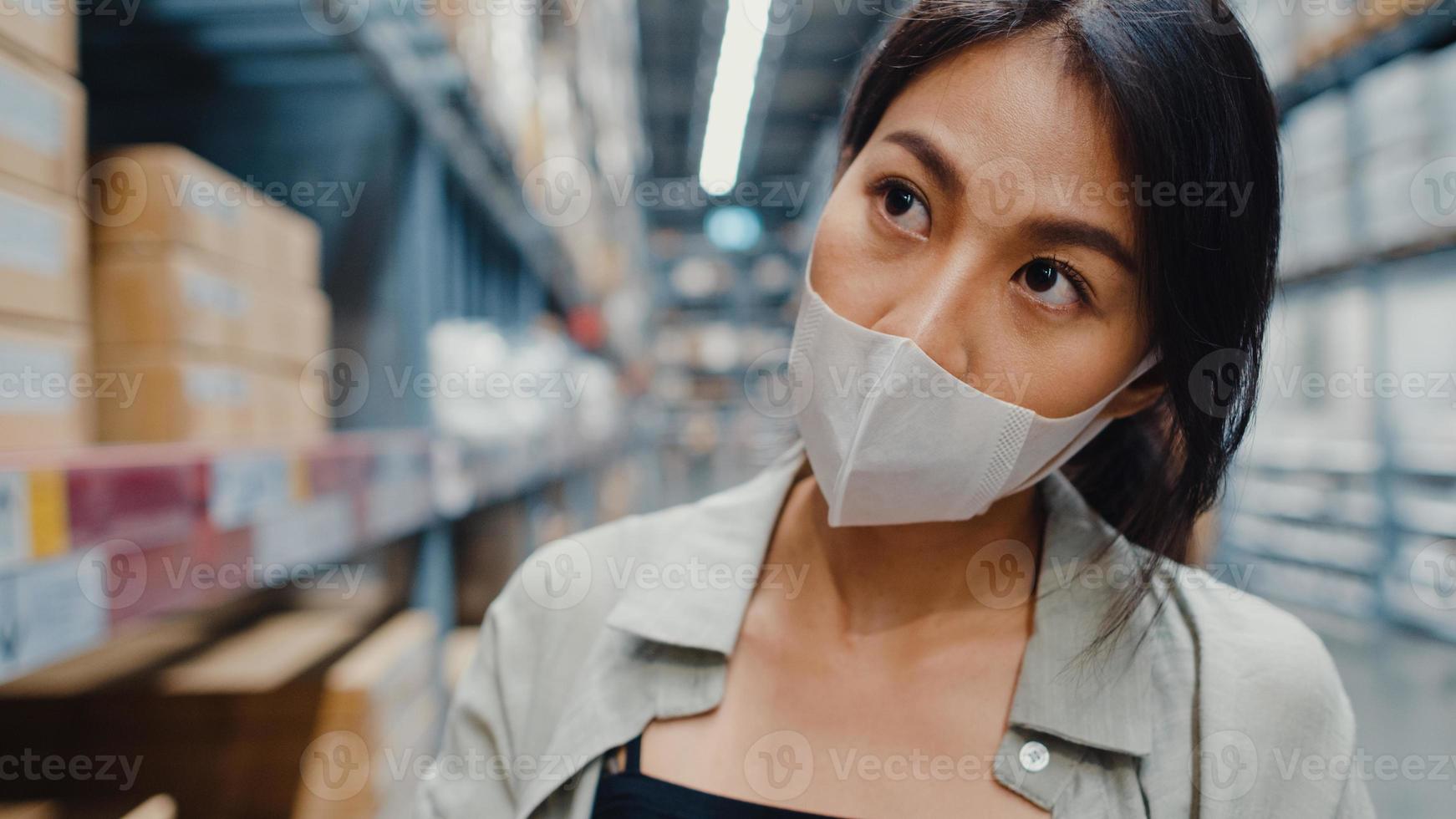 Young Asia businesswoman manager wear face mask looking for goods using digital tablet checking inventory levels stand in retail shopping center. Distribution, Logistics, Packages ready for shipment. photo