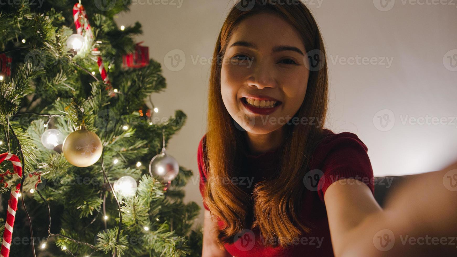 Mujer joven de Asia con videollamada de teléfono inteligente hablando con pareja, árbol de Navidad decorado con adornos en la sala de estar en casa. distanciamiento social, noche de navidad y fiesta de fin de año. foto