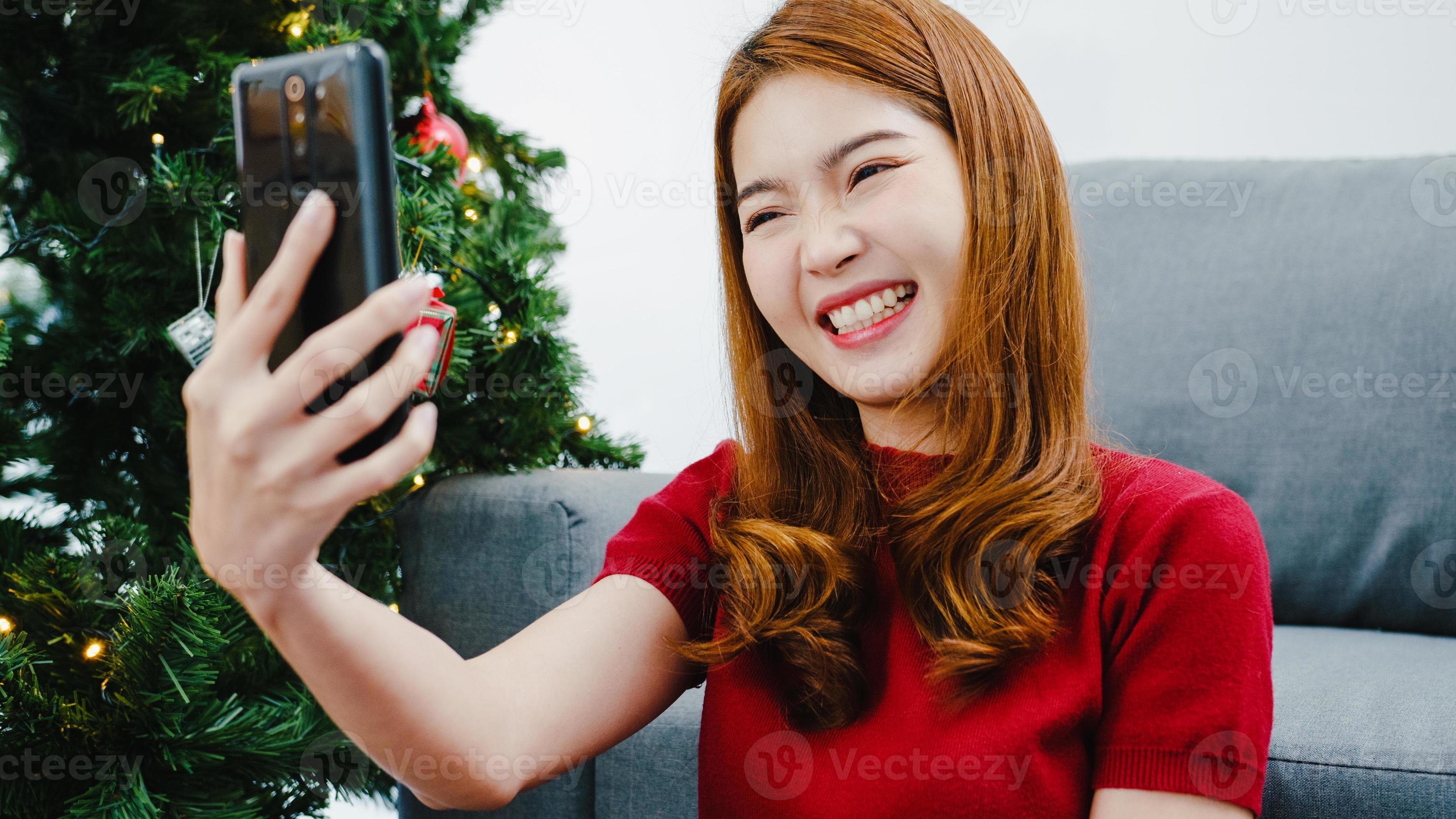 Young Asian female using smart phone video call talking with couple with XMas present box, Christmas tree decorated with ornament in living room at home photo
