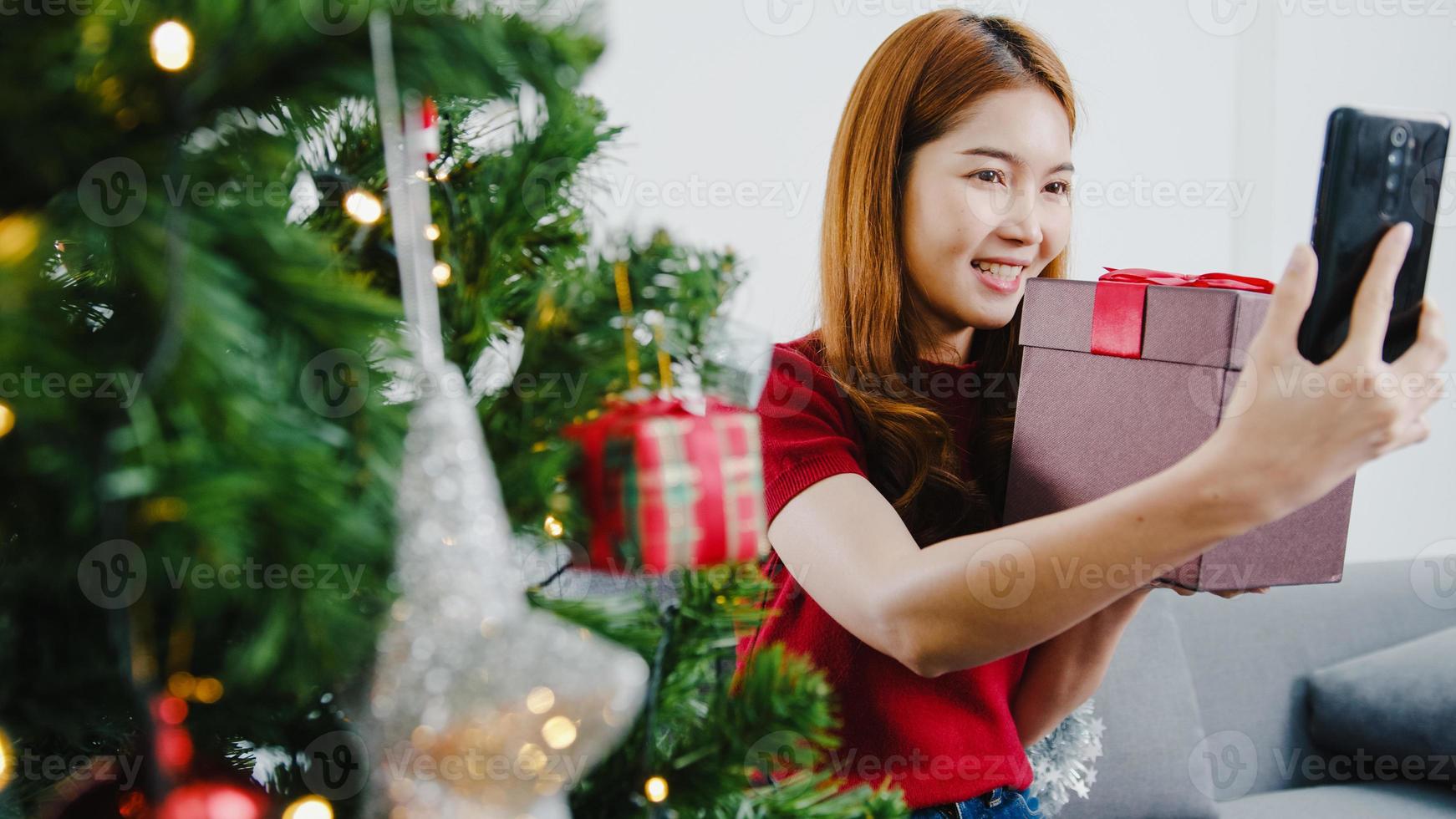 Young Asian female using smart phone video call talking with couple with X'Mas present box, Christmas tree decorated with ornament in living room at home. Christmas and New Year holiday festival. photo