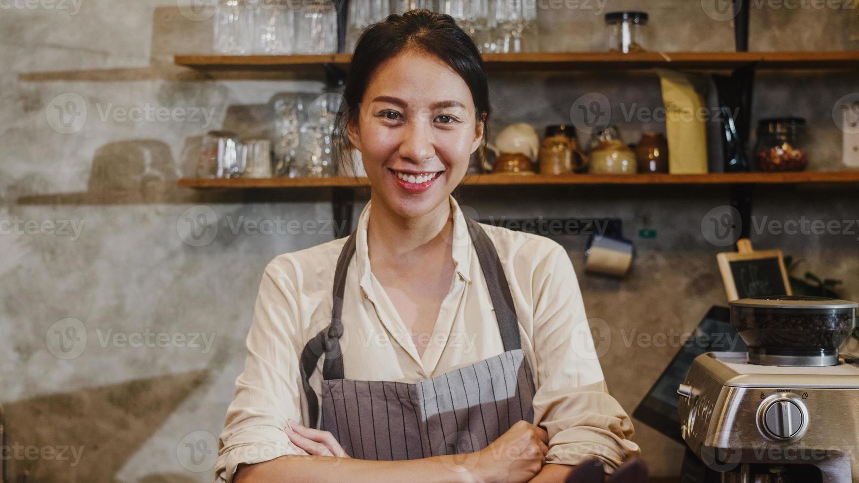 retrato joven mujer asiática barista sentirse feliz sonriendo en el café urbano. propietario de una pequeña empresa chica indonesia en delantal relajarse con una sonrisa con dientes mirando a la cámara de pie en el mostrador de la cafetería. foto