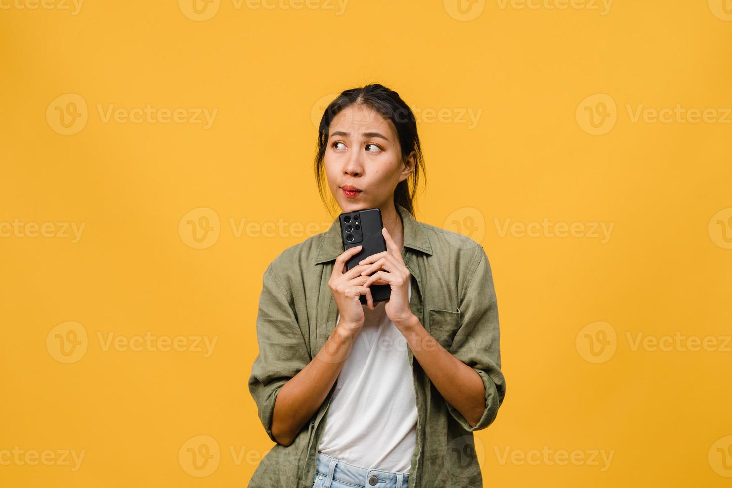 Pensando soñando a una joven asiática que usa el teléfono con expresión positiva, vestida con un paño casual sintiendo felicidad y de pie aislado sobre fondo amarillo. feliz adorable mujer alegre se regocija con el éxito. foto