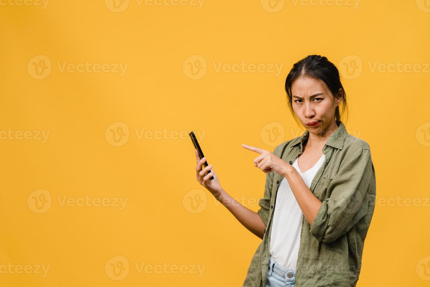 Thinking dreaming young Asia lady using phone with positive expression, dressed in casual cloth feeling happiness and stand isolated on yellow background. Happy adorable glad woman rejoices success. photo