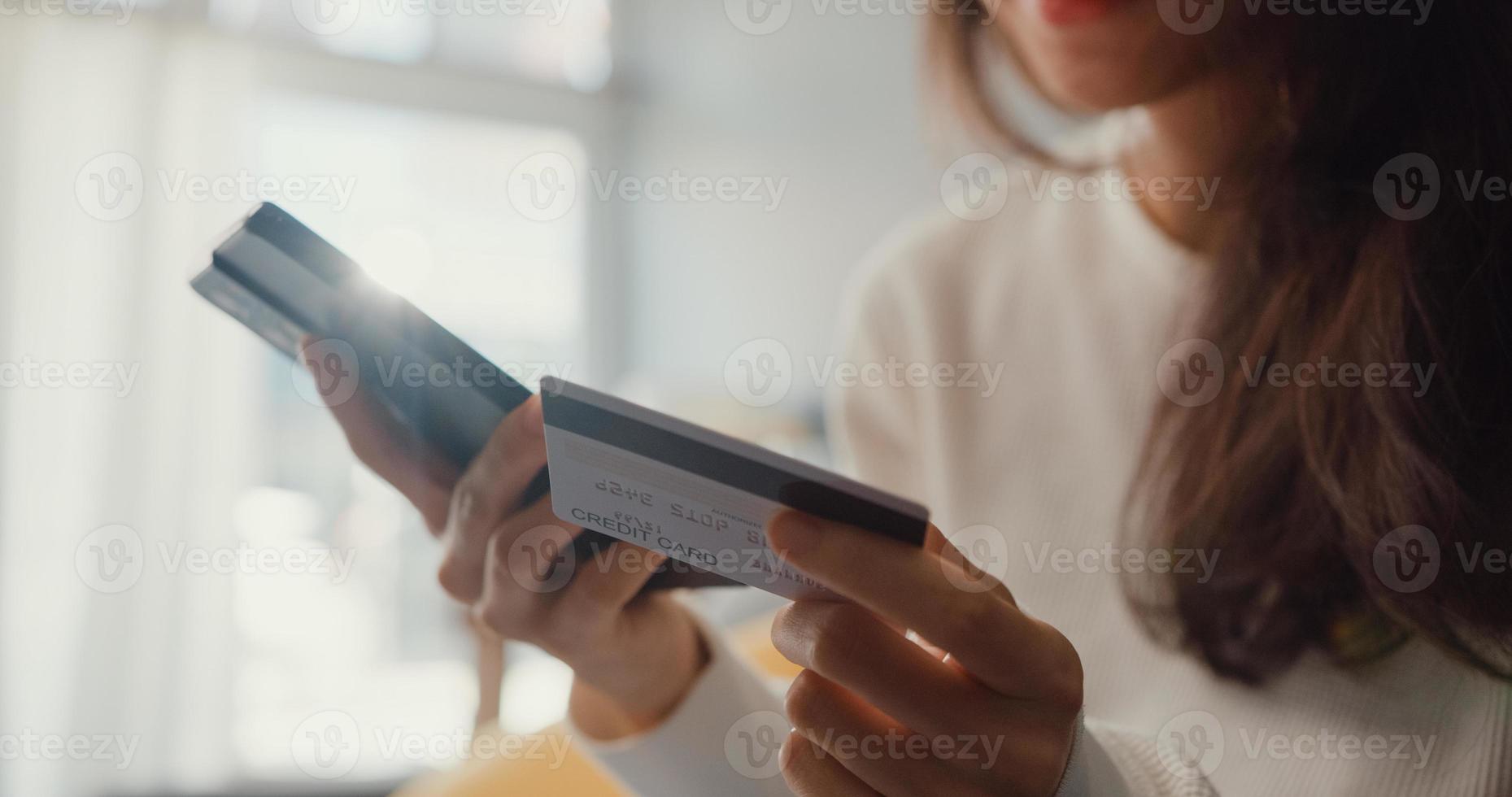 Closeup joven mujer asiática usa teléfono celular, compra productos en línea y paga la factura con tarjeta de crédito, se sienta en el sofá en la sala de estar de la casa, actividad de cuarentena, actividad divertida para la prevención del coronavirus. foto