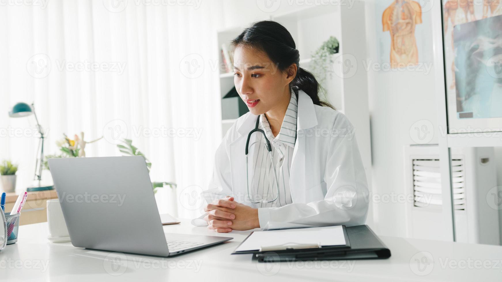 Joven doctora asiática en uniforme médico blanco con estetoscopio usando computadora portátil hablando por videoconferencia con el paciente en el escritorio en la clínica de salud u hospital. concepto de consulta y terapia. foto