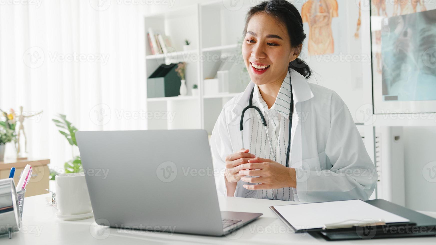 Young Asia lady doctor in white medical uniform with stethoscope using computer laptop talking video conference call with patient at desk in health clinic or hospital. Consulting and therapy concept. photo