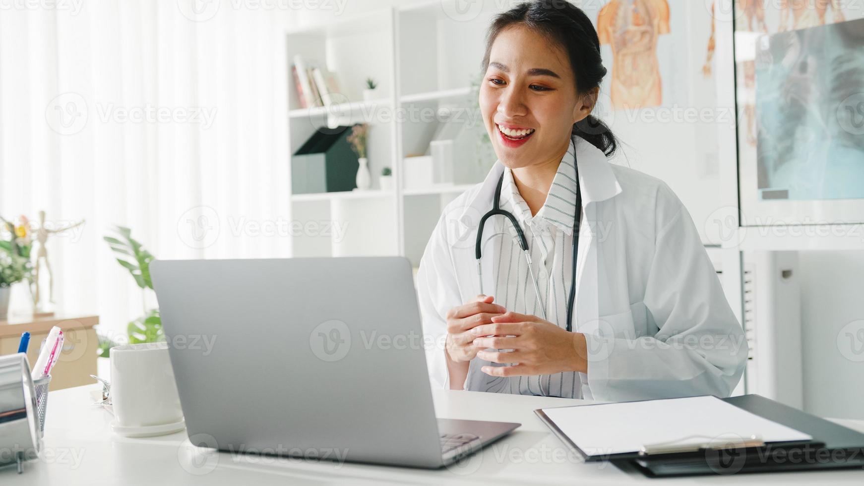 Joven doctora asiática en uniforme médico blanco con estetoscopio usando computadora portátil hablando por videoconferencia con el paciente en el escritorio en la clínica de salud u hospital. concepto de consulta y terapia. foto