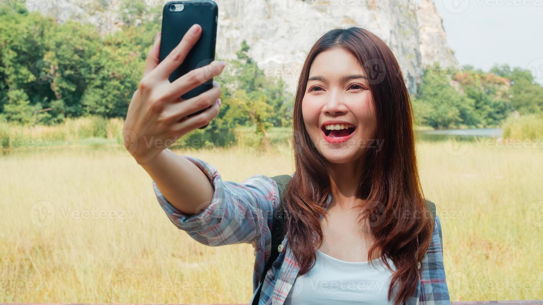Cheerful young traveler Asian lady with backpack recording video vlog live streaming on phone upload in social media at mountain lake. Korean girl happy enjoy holiday trip. Lifestyle travel and relax. photo