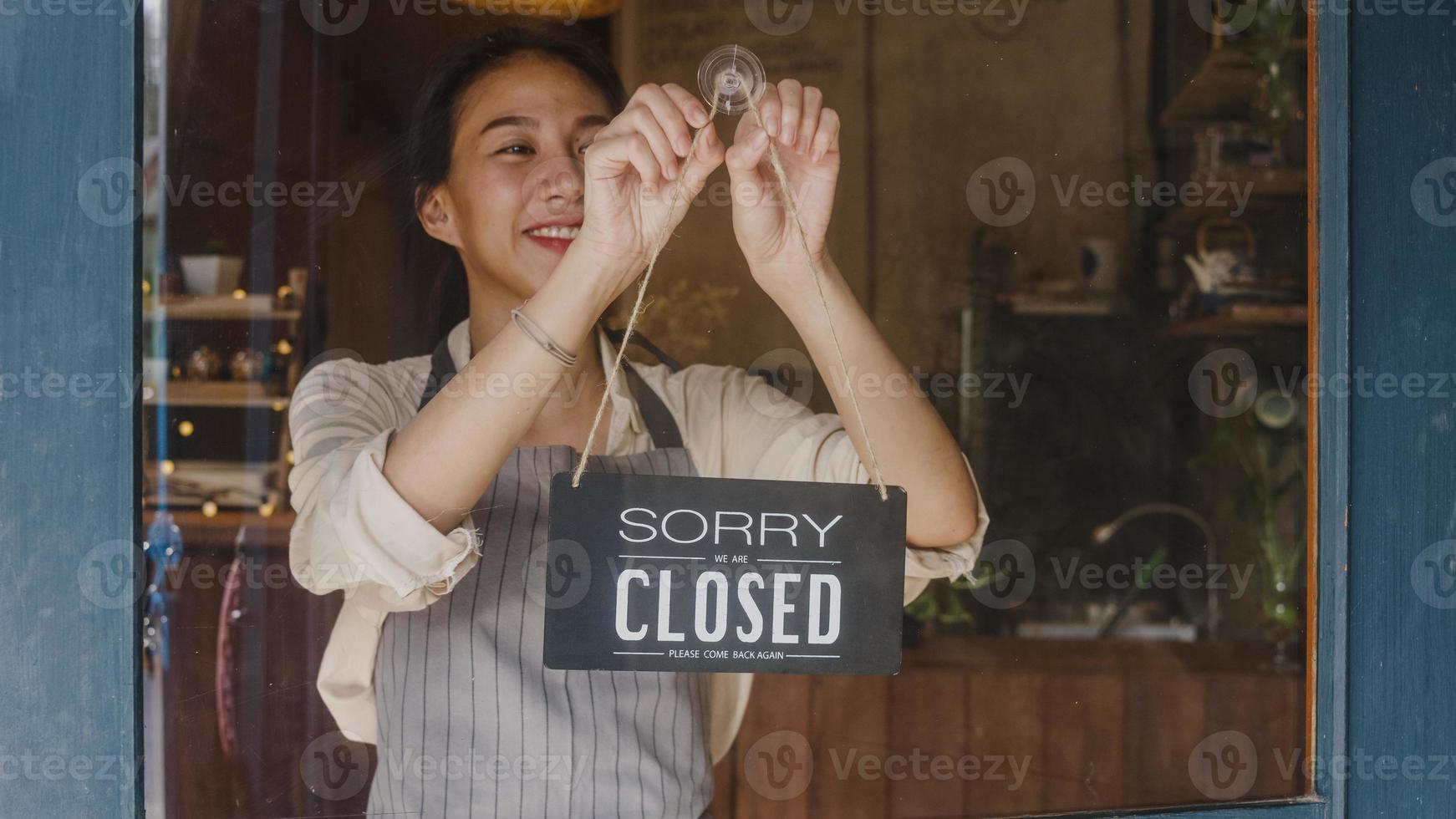 Young Asia manager girl changing a sign from closed to open sign on door cafe looking outside waiting for clients after lockdown. Owner small business, food and drink, business reopen again concept. photo