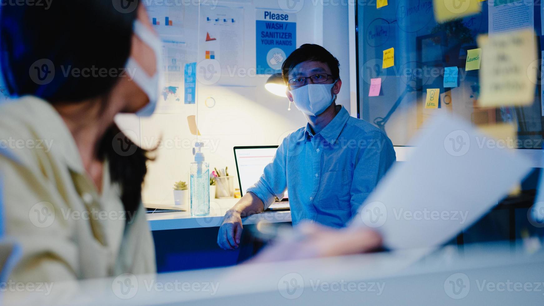 Feliz empresaria asiática usa mascarilla para el distanciamiento social en una nueva situación normal para la prevención de virus mientras se discute una reunión de lluvia de ideas de negocios juntos compartir datos en el trabajo en la noche de la oficina foto