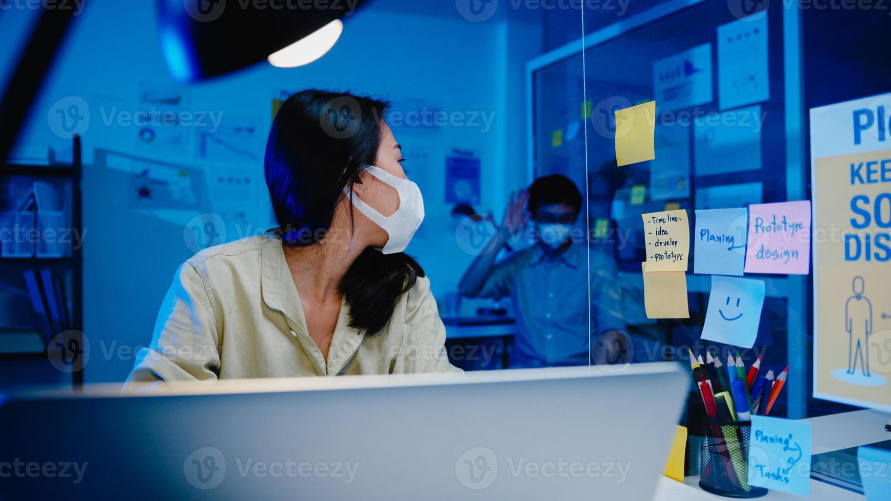 Asian businessman turn off computer and say goodbye his colleague who still working when he get off work after finish working overtime in small modern home office night. Coworker partnership concept. photo