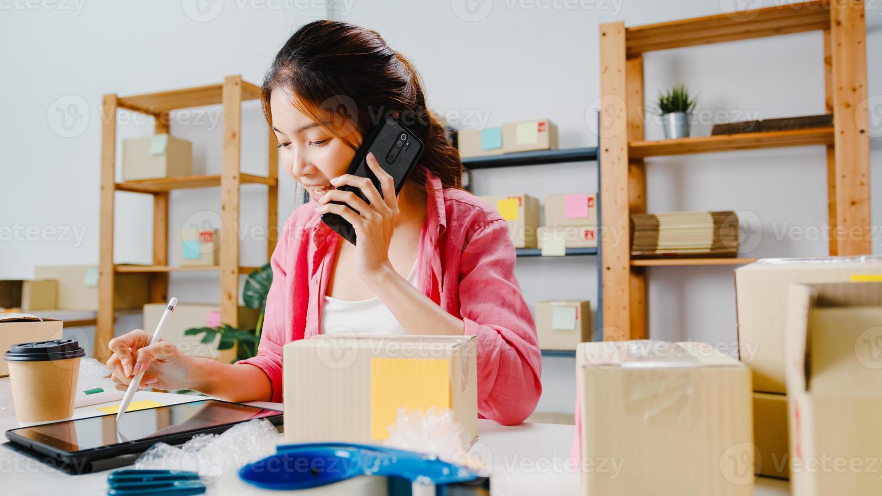 Young Asia businesswoman using mobile phone call receiving purchase order and check product on stock, work at home office. Small business owner, online market delivery, lifestyle freelance concept. photo