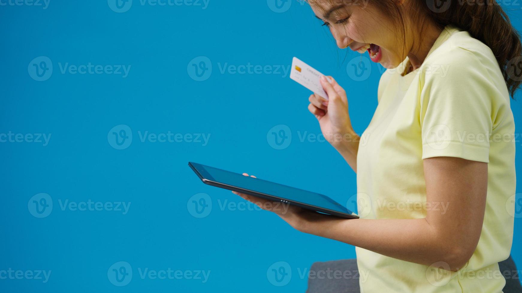 Jovencita asiática usando tableta y tarjeta bancaria de crédito con expresión positiva, sonrisa amplia, vestida con ropa casual y parada aislada sobre fondo azul. feliz adorable mujer alegre se regocija con el éxito. foto