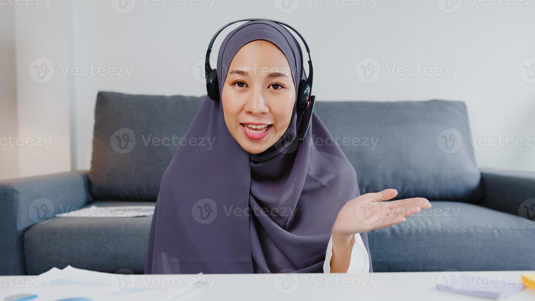 Asia muslim lady wear headphone using computer laptop talk to colleagues about plan in video call meeting while remotely work from home at living room. Social distancing, quarantine for corona virus. photo