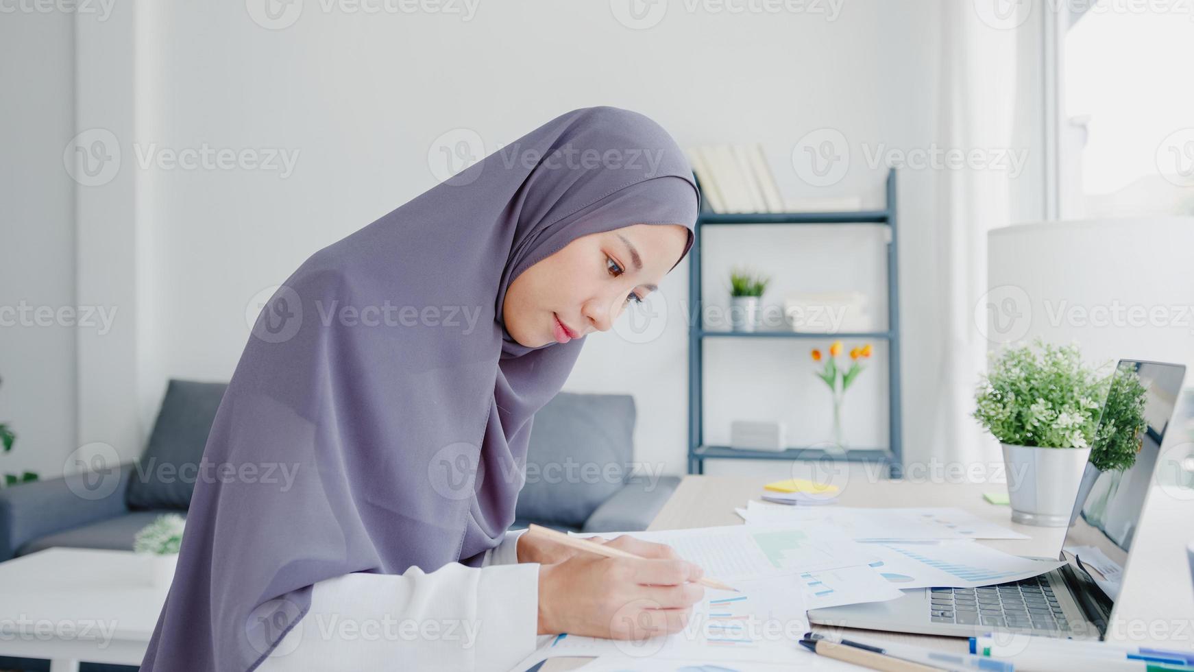 Beautiful Asia muslim lady in headscarf casual wear using laptop in living room at house. Remotely working from home, new normal lifestyle, social distancing, quarantine for corona virus prevention. photo